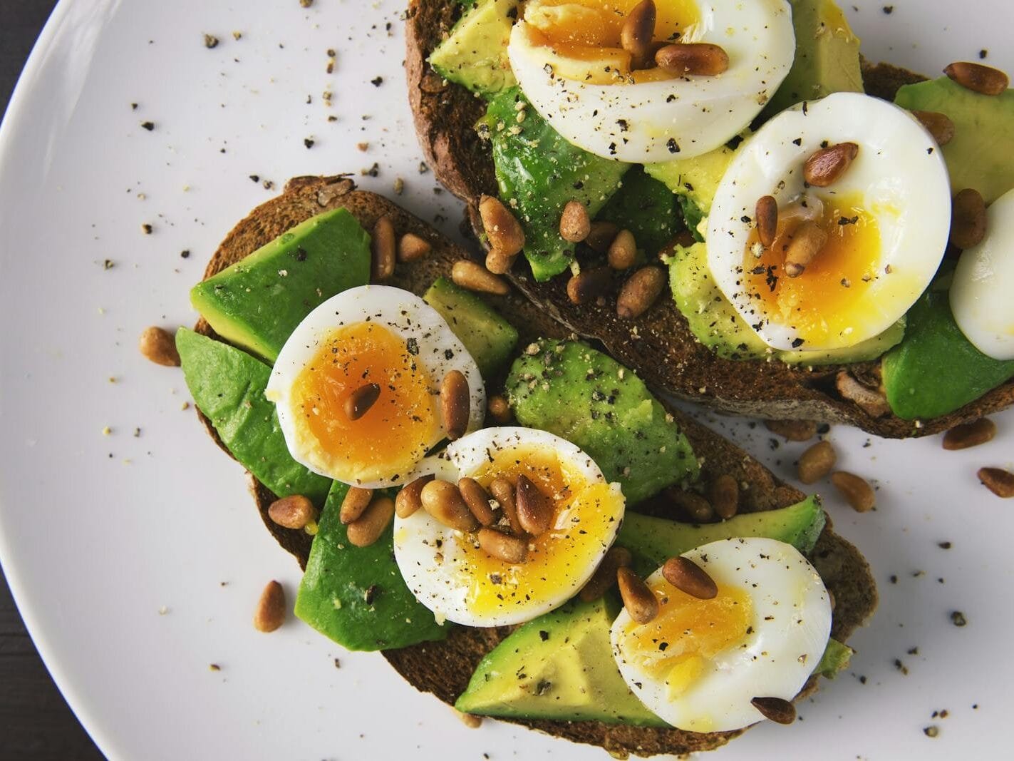 Delicious avocado toast topped with soft-boiled eggs and pine nuts on a white plate.