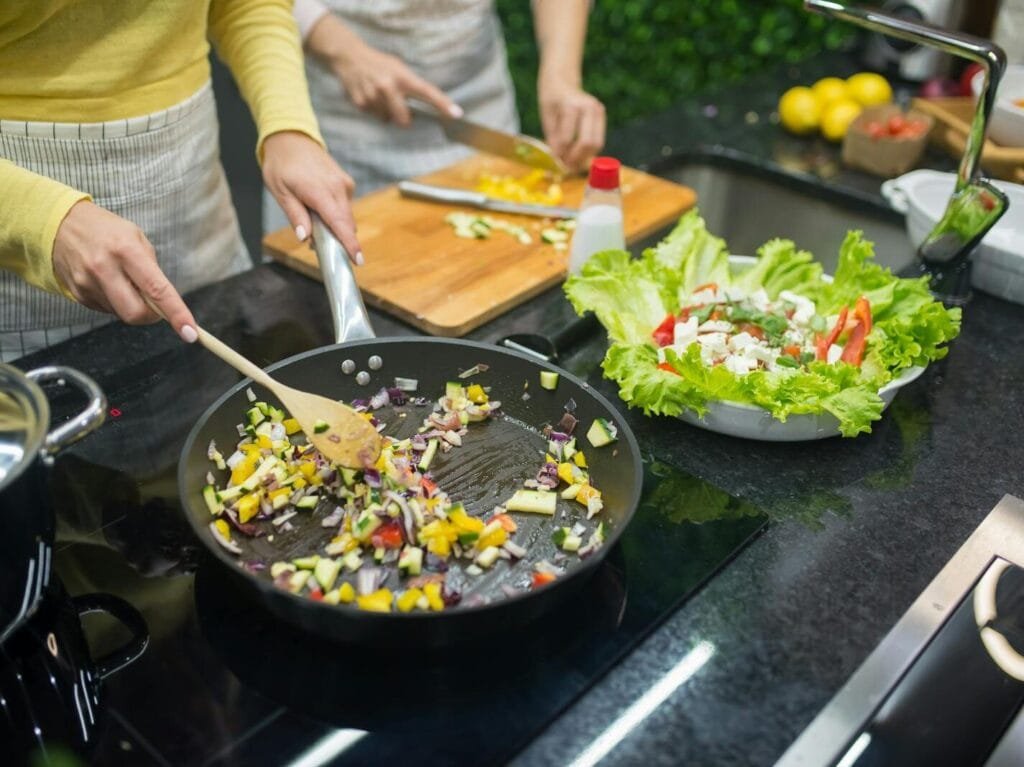 Two adults cooking fresh vegetables on a stove, preparing a healthy meal in a modern kitchen.